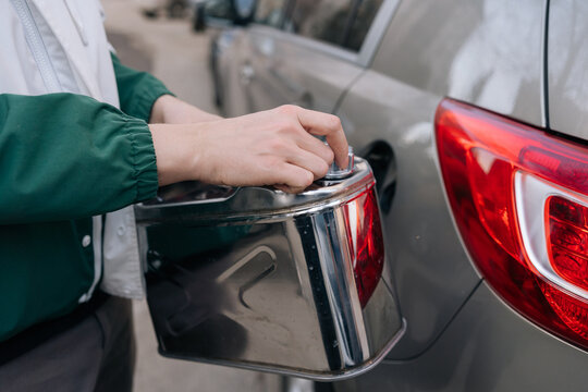 Person filling vehicle fuel tank with gasoline from silver metal jerrican, addressing themes of energy crisis, rising fuel prices, car dependency.