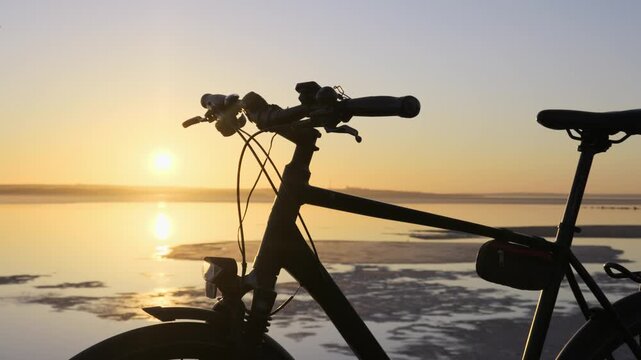 A commuter bicycle stands in silhouette against a beautiful golden hour sunset over calm water. Concept of outdoor recreation, peaceful travel, active lifestyle, and solitary evening rides.