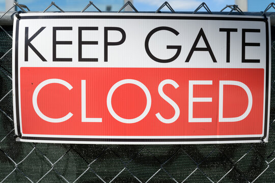 Keep Gate Closed sign with white-over-red split, sans-serif type, and black border, tied to a chain link fence