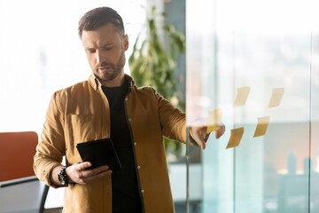 Fototapeta premium Focused Caucasian Male Professional Analyzing Data On Tablet While Standing In Modern Office Environment, Engaged In Planning And Strategy Discussion. Business Concept.