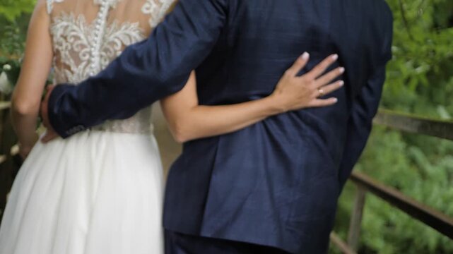 A bride in an ornate white dress and a groom in a blue suit wrap their arms around each other while walking away on a rustic bridge surrounded by green foliage.