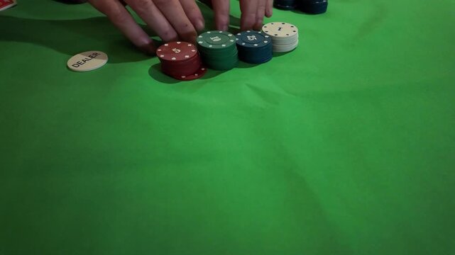 A top-down, close-up shot of a person's hands placing casino chips on a vibrant green table. The chips are stacked in different colors and denominations, depicting a moment of play or betting. The