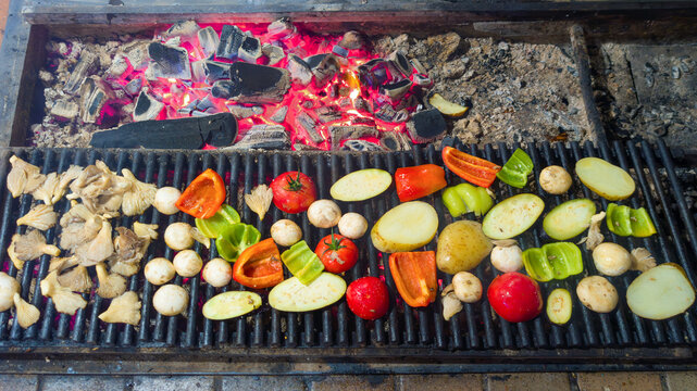 Colorful selection of vegetables including bell peppers, tomatoes, potatoes, and various mushrooms cooking on a hot grill over burning coals, preparing a delicious meal