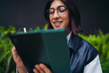 Close up portrait of smiling hispanic woman looking down at tablet. Beautiful spanish female student with eyeglasses typing text chat layout message near blurred city park background. © BullRun