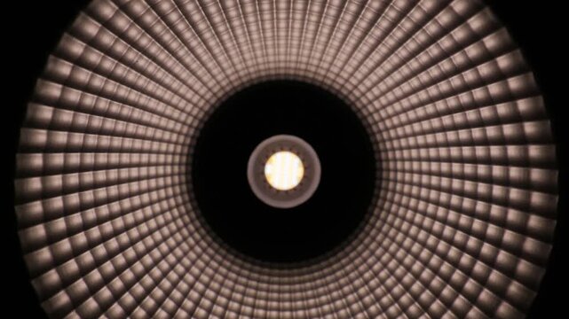 Illuminated LED light bulb glows brightly in the center of a dark frame. Concentric circular rings create a textured tunnel pattern around the light. Individual diode dots are clearly visible.