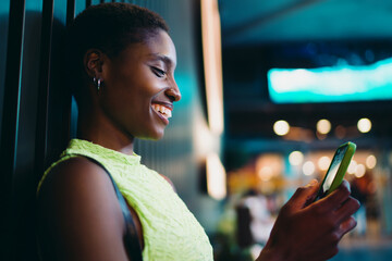 Profile portrait of laughing African American woman looking at green smartphone screen indoors. Joyful dark skinned female youth reading comments or text messaging friends near wall. © BullRun