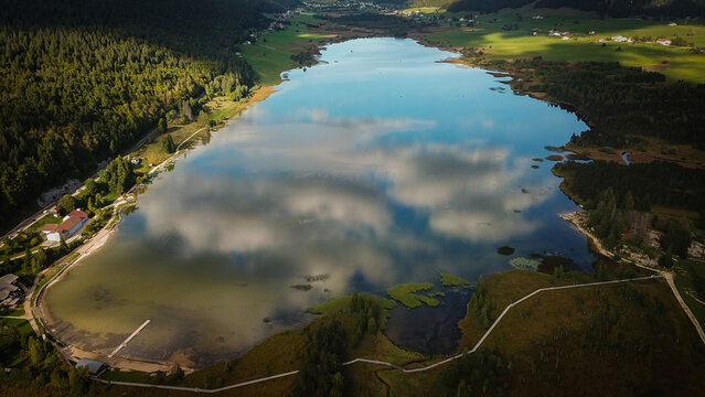 Aerial view of Lac des Rousses surrounded by lush green forests, rolling hills, and a wooden walking path along the shoreline in Les Rousses, Bourgogne-Franche-Comte, France.