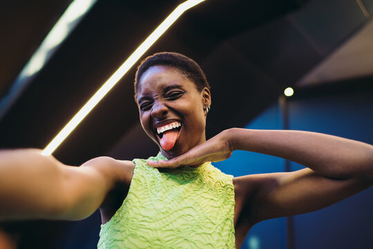 Excited black woman in neon dress showing tongue and capturing funny selfie on mobile phone. Cheerful African American youth posing for social media publication under terminal lights.