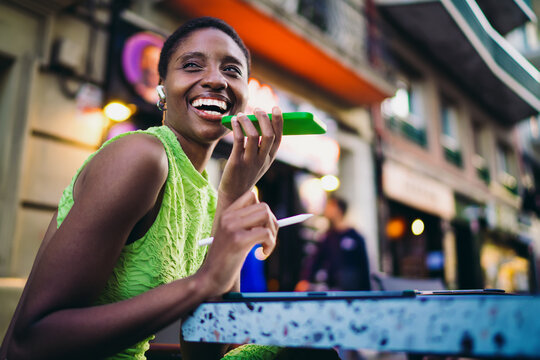 Overjoyed African American woman laughing out loud while holding smartphone near mouth for voice command. Happy dark skinned female youth enjoying active mobile conversation at street cafe.