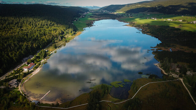 Aerial view of Lac des Rousses surrounded by lush green forests and rolling hills under a sky with scattered clouds in Les Rousses, Bourgogne-Franche-Comte, France.