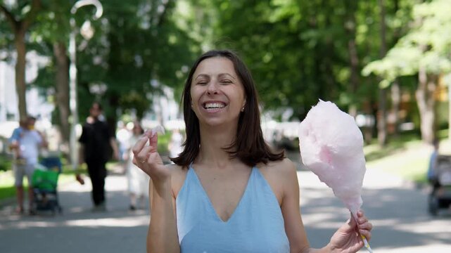 A joyful beautiful woman eats cotton candy in a portrait park in summer 4k
