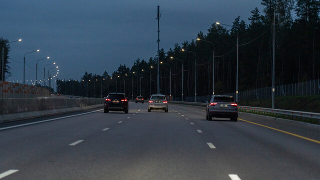 Darkening sky lines a multi-lane highway at dusk. Taillights of several cars glow as they move toward the viewer. Streetlights illuminate the road, flanked by trees