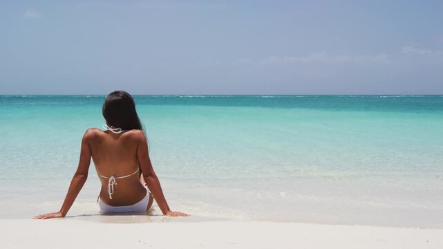 Travel vacation girl in white bikini relaxing in tropical beach sun tanning in summertime.W oman in relaxation mode at idyllic stunning tropical luxury resort hotel. SLOW MOTION.