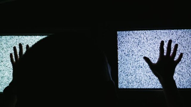 Silhouette of a person stands in a dark room with hands raised toward two glowing screens filled with analog television noise. Concept of media overload, psychological distress, and horror.
