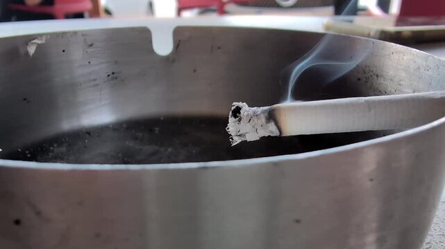 Close up of burning cigarette resting on a metal ashtray, slowly releasing toxic smoke, symbolizing addiction, unhealthy habit, and the risks of tobacco consumption