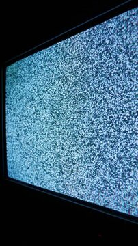 Vertical television monitor glows brightly with textured white noise static in a dark room. The screen displays a random pattern of cyan and blue pixels indicating no signal.