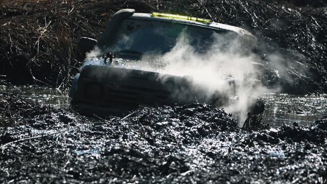 Rugged offroad vehicle struggles through deep, dark mud. Steam rises from hot engine during intense motorsport competition. Backlit scene creates dramatic, high-contrast look for powerful adventure