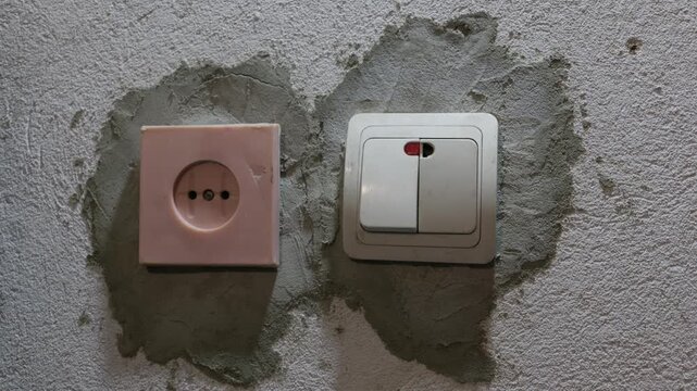 A hand presses a white double light switch beside an old electrical outlet on a freshly plastered wall. Concept of home renovation, interior construction, and electrical upgrades.