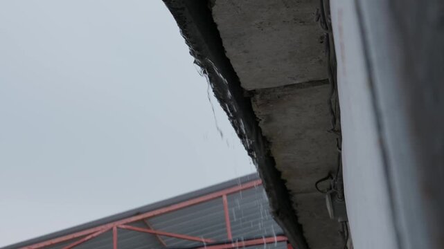 Rain water drips steadily from a worn concrete building gutter under a gloomy overcast sky during a cold wet day. Concept of rainy weather, building maintenance, and seasonal change.