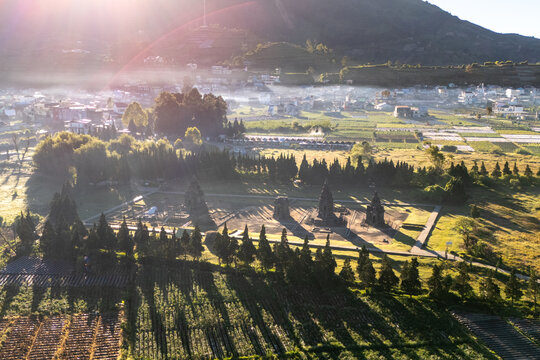 Aerial view of the ancient Arjuna Temple complex surrounded by lush agricultural fields, cypress trees, and morning mist under the golden sunrise light in the Dieng Plateau, Indonesia.