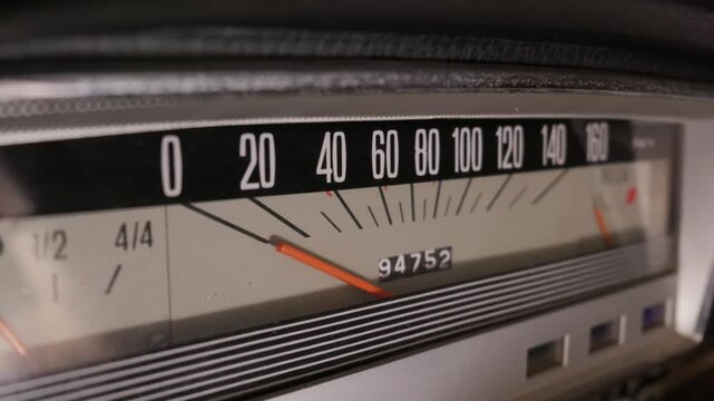 A macro view reveals the dashboard details of a classic vintage automobile. The analog linear speedometer, odometer digits, and fuel gauge feature striking orange indicator needles.
