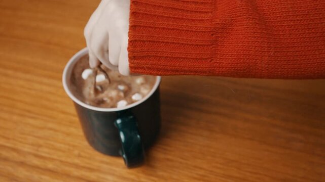 Top angle shot of a woman in red sweater stirring hot chocolate with marshmallows, Close up of female hand using a cat shaped spoon to mix hot cocoa mug on wooden table