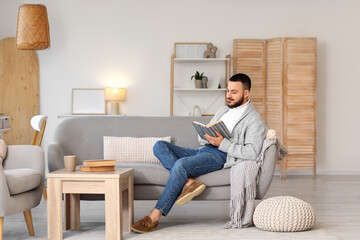 Fototapeta premium Handsome young man reading book on grey sofa at home