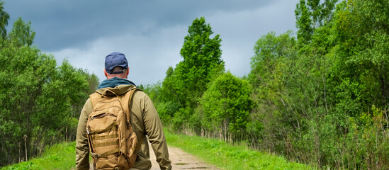 Tourist with backpack stands at beginning of forest road with back to camera and prepares to continue independent journey © Dmitrii Masquerade