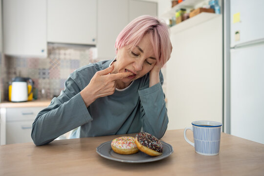 Doubtful mature woman with dieting problems and sugar addiction tasting sweet pastry, female with guilty expression and low willpower licking finger in glaze, self hesitation and mental breakdown.