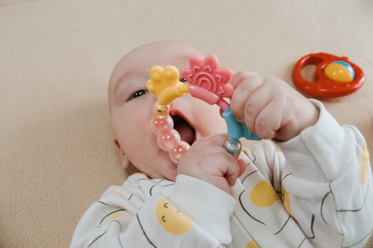 Baby lying on bed and gripping teething ring with both hands. Child chewing toy for teething relief. Exploring textures while soothing sore gums. Sensory discovery at home.