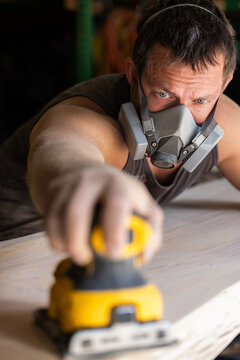 Man wears protective mask while sanding wood with an orbital sander. Focused expression shows care and precision in work.