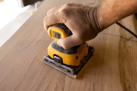Hand guides power orbital sander over wooden surface. Workbench hints at craftsmanship or DIY project