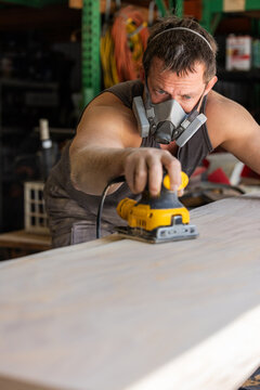Man wears protective mask while sanding wood with an orbital sander. Focused expression shows care and precision in work.