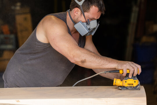 Man wears protective mask while sanding wood with an orbital sander. Focused expression shows care and precision in work.