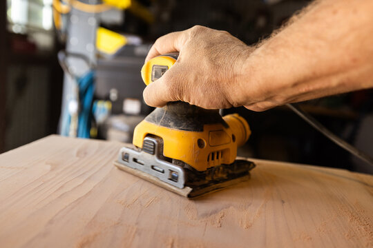 Hand guides power orbital sander over wooden surface. Workbench hints at craftsmanship or DIY project
