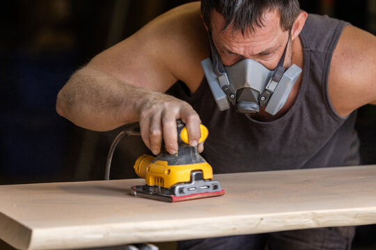 Man wears protective mask while sanding wood with an orbital sander. Focused expression shows care and precision in work.