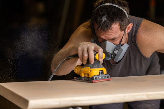 Man wears protective mask while sanding wood with an orbital sander. Focused expression shows care and precision in work.
