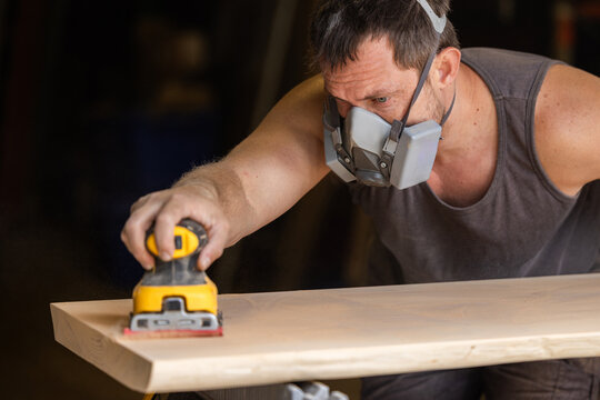 Man wears protective mask while sanding wood with an orbital sander. Focused expression shows care and precision in work.
