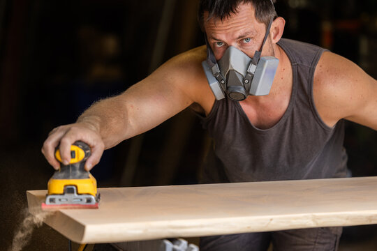 Man wears protective mask while sanding wood with an orbital sander. Focused expression shows care and precision in work.