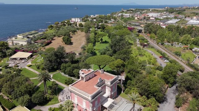 Panoramic view of Villa Guarracino pink historic mansion with formal gardens, manicured lawns and palm trees overlooking the Tyrrhenian sea, Torre Annunziata, Naples, Italy
