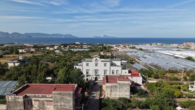 Aerial drone view of white historic villa among rooftops with coastal greenhouses and sea with islands in background, Torre del Greco, Italy
