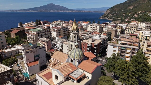 Aerial drone view of Vico Equense town with Chiesa dei Santi Ciro e Giovanni dome, terracotta rooftops and Mount Vesuvius across the gulf, Italy

