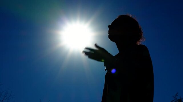 Woman suffers from the heat. A silhouette of a woman against a blue sky and hot sun fans her face with her hands, suffering from the heat.