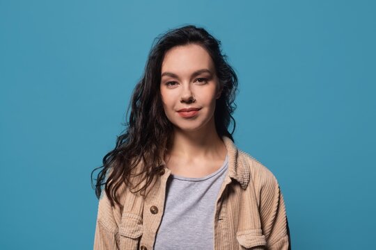 Smiling Young Woman With Long Dark Hair, Standing Confidently In Studio With Solid Blue Background. She Poses For Camera In Professional Setting, Reflecting Positivity And Strength. Business Concept.