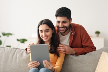 Obraz premium Happy Young Couple, Asian Male And Female, Enjoying Time Together While Using Tablet On Couch At Home. Smiling Faces Indicate Joyful Moment With Technology In Cozy Living Room. Lifestyle Concept.