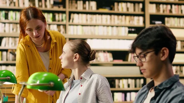 Tutor guiding seated student in library and helping with coursework, creating collaborative learning academic study session, Mentor in yellow shirt pointing toward laptop, tutoring.