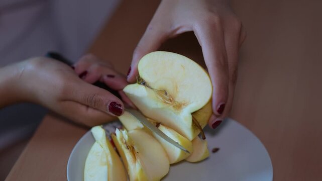 Feminine hands with red nail polish carefully cutting a fresh yellow apple into thin slices with a kitchen knife and placing them on a plate, preparing a healthy organic fruit snack