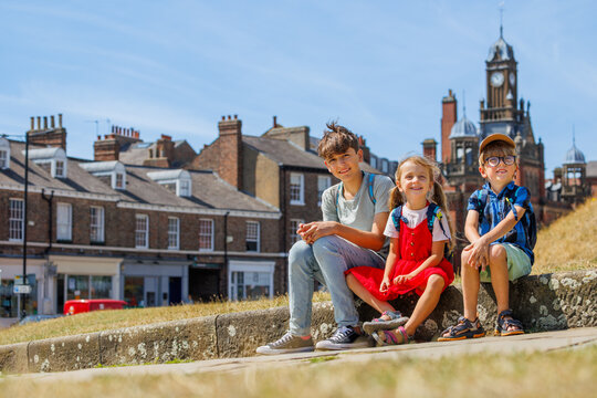 Three children sit on sunny town ledge with backpacks