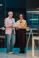 Female it professionals collaborating in a data center, analyzing information on a clipboard with...
