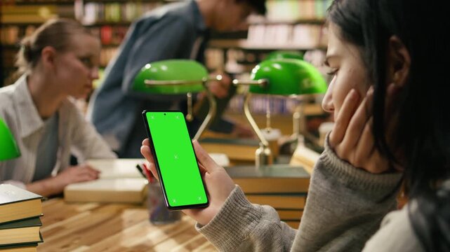 Vertical chroma key editable screen on mobile phone held by student during library study group at desk, providing replaceable screen space with peers blurred in background, supporting green screen.